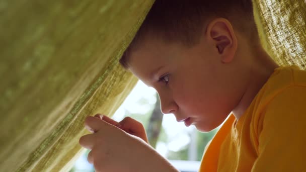 Jongen alleen in het huis met een deken spelen van een computerspel op een smartphone. Technologie generatie. Kleuterscholier brengt zomertijd door in tent met mobiele apps. — Stockvideo