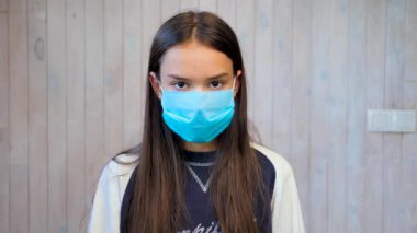 Tıbbi koruyucu maske takan genç kız kameraya bakıyor. Covid-19, koronavirüs salgını. Açık gri duvar arka planında tıbbi koruyucu maskeli güzel esmer kız portresi..