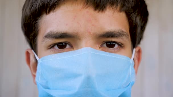 Blisko twarzy młodego człowieka w niebieskiej masce medycznej i patrzącego w kamerę. Portret pacjenta w masce ochronnej, chłopiec zapobiegający pandemii grypy koronawirusowej w pomieszczeniach — Wideo stockowe