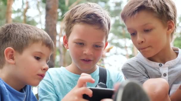 Děti se dívají na obrazovku. Děti hrají videohry venku. Tři evropské děti sledují hru, hrají si na mobilu v pozadí parku. Chlapec s pihami na obličeji pohled na mobilní — Stock video