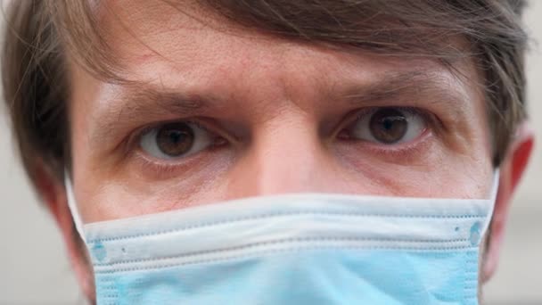 Vista de primer plano de un hombre caucásico con una máscara facial durante la pandemia del Coronavirus Covid-19. Tipo con máscara médica a prueba de virus en la cara, mirando a la cámara mientras está aislado en casa. — Vídeos de Stock