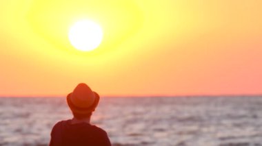 Şapkalı genç adam okyanusun kenarında gün batımını seyrediyor. Erkek gezgin doğu ve dalgaların manzarasının tadını çıkarıyor. Güzel bir akşamda denize bakan bir adamın silüeti. Tatil kavramı