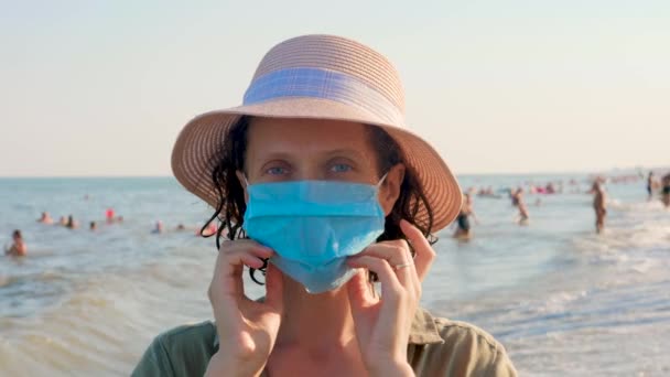 Urlaub und Coronavirus-Ausbruch. Frau mit Einwegmaske blickt in die Kamera. Meereshintergrund mit Reisenden in Gesichtsmaske im Vordergrund. Vorbeugung gegen Coronavirus. Sichere und glückliche Dame in Maske — Stockvideo