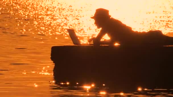 Фрілансер, який друкує на ноутбуці над заходом сонця, відображається у морській воді. Чоловік, блогер з ноутбуком на пірсі. Віддалена робота, концепція свободи. Схід сонця, що відбивається у воді . — стокове відео