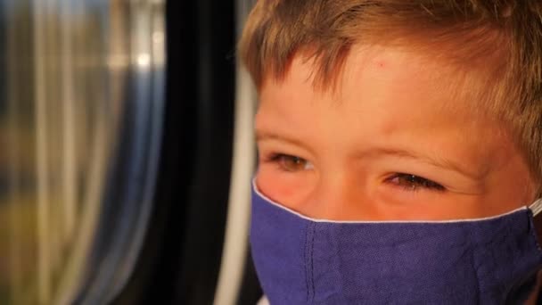 Chico mirando por la ventana durante el viaje en tren. Primer plano de niño en máscara de algodón. Luz solar en la cara del pasajero. Niño con máscara protectora en el transporte. Emociones faciales de viajero pequeño — Vídeo de stock