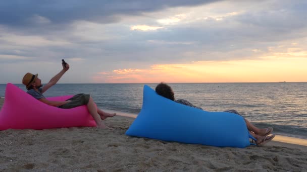 Gün doğumunda plajda şişme koltukta oturan çift. Genç adam sahilde gün batımında selfie çekiyor. Erkek ve kadın tatilde dinleniyor. Psikoloji, ilişki kavramı. — Stok video