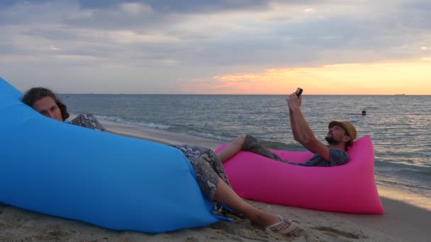 Пара сидить на ліжку на пляжі під час сходу сонця. Чоловік і жінка відпочивають у відпустці. Психологія, концепція стосунків. Стильний чоловік робить селфі на надувному дивані на фоні заходу сонця — стокове відео