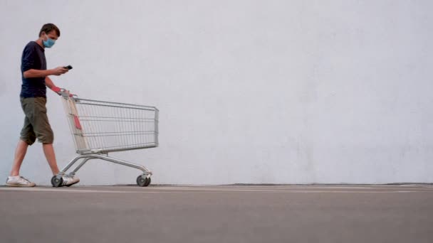 Hombre en máscara con Smartphone empujando el carro de la compra. Cliente con carrito de compras a lo largo de la pared de la tienda. Guy en una mascarilla desechable después de la muerte se sumergió en la navegación por Internet — Vídeos de Stock