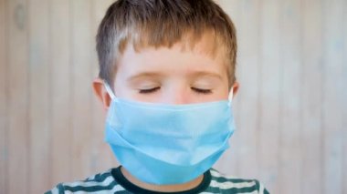 Tıbbi bir maske takmış küçük bir çocuk kameraya bakıyor ve gülümsüyor. Kahverengi Gözlü Küçük Çocuklar Koruyucu Maskeli Cam için poz veriyorlar. Çocuk Duyguları 'ndan. Coronavirus Covid-19. Kapalı