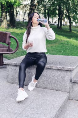Güzel esmer kadın, parkta korkulukta otururken bir şişeden su içer ve gözlerini kapatır.