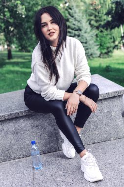 Güzel esmer, egzersizden sonra dinleniyor. Parktaki mermer korkulukta oturuyor.