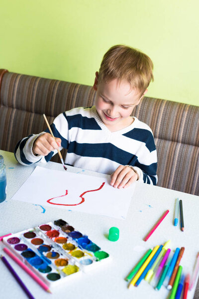 Smiling and happy caucasian child with blond hair is diligently drawing a human with watercolours indoors vertically
