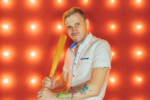 A man with a baseball bat in his hands posing on a red background of spotlights. Guy with a tattoo on his arm on a red background.