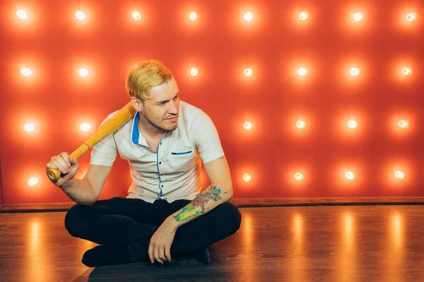 A man with a baseball bat in his hands posing on a red background of spotlights. Guy with a tattoo on his arm on a red background.