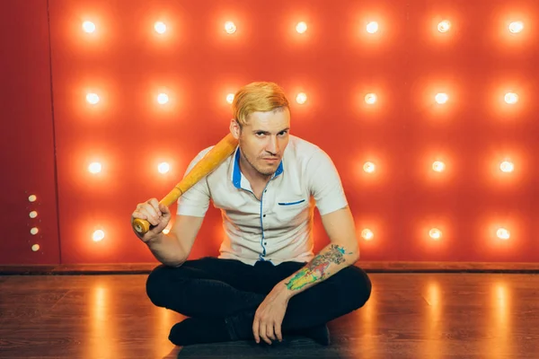A man with a baseball bat in his hands posing on a red background of spotlights. Guy with a tattoo on his arm on a red background.