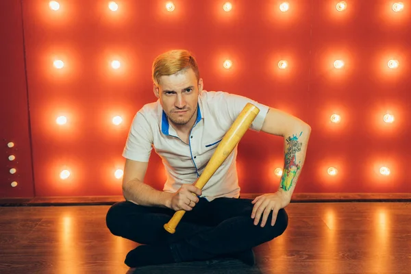 A man with a baseball bat in his hands posing on a red background of spotlights. Guy with a tattoo on his arm on a red background.