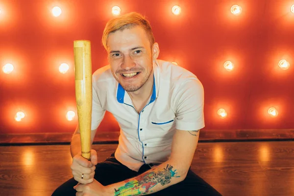 A man with a baseball bat in his hands posing on a red background of spotlights. Guy with a tattoo on his arm on a red background.