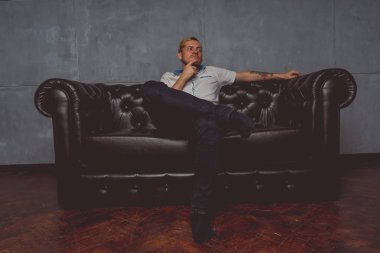 dövme deri kanepede poz elinde olan genç bir adam. sokak tarzı kıyafetler: beyaz gömlek ve siyah kot. kısa saç ve temiz cilt. duygusal bir öğrenci portresi