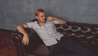 dövme deri kanepede poz elinde olan genç bir adam. sokak tarzı kıyafetler: beyaz gömlek ve siyah kot. kısa saç ve temiz cilt. duygusal bir öğrenci portresi