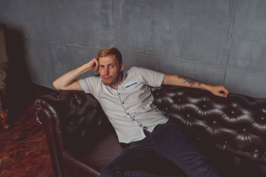 dövme deri kanepede poz elinde olan genç bir adam. sokak tarzı kıyafetler: beyaz gömlek ve siyah kot. kısa saç ve temiz cilt. duygusal bir öğrenci portresi