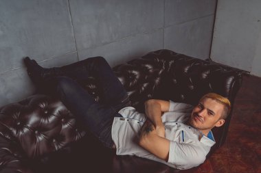 dövme deri kanepede poz elinde olan genç bir adam. sokak tarzı kıyafetler: beyaz gömlek ve siyah kot. kısa saç ve temiz cilt. duygusal bir öğrenci portresi