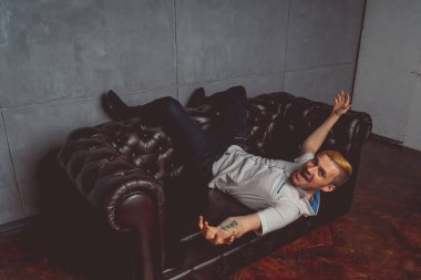 dövme deri kanepede poz elinde olan genç bir adam. sokak tarzı kıyafetler: beyaz gömlek ve siyah kot. kısa saç ve temiz cilt. duygusal bir öğrenci portresi