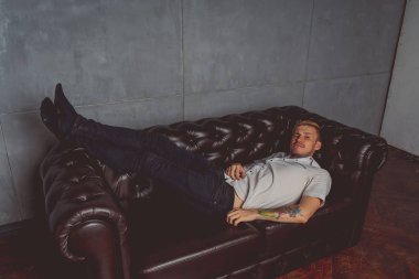 dövme deri kanepede poz elinde olan genç bir adam. sokak tarzı kıyafetler: beyaz gömlek ve siyah kot. kısa saç ve temiz cilt. duygusal bir öğrenci portresi