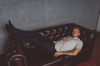 dövme deri kanepede poz elinde olan genç bir adam. sokak tarzı kıyafetler: beyaz gömlek ve siyah kot. kısa saç ve temiz cilt. duygusal bir öğrenci portresi