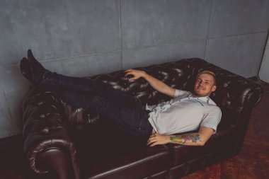 dövme deri kanepede poz elinde olan genç bir adam. sokak tarzı kıyafetler: beyaz gömlek ve siyah kot. kısa saç ve temiz cilt. duygusal bir öğrenci portresi