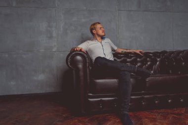 dövme deri kanepede poz elinde olan genç bir adam. sokak tarzı kıyafetler: beyaz gömlek ve siyah kot. kısa saç ve temiz cilt. duygusal bir öğrenci portresi