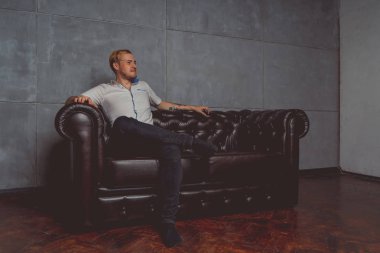 dövme deri kanepede poz elinde olan genç bir adam. sokak tarzı kıyafetler: beyaz gömlek ve siyah kot. kısa saç ve temiz cilt. duygusal bir öğrenci portresi