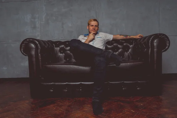 a young man with a tattoo on his hand posing on a leather couch. street style clothes: white shirt and black jeans. short hair and clean skin. emotional portrait of a student