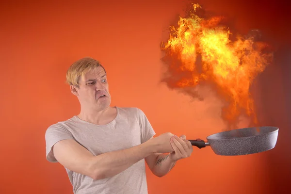 Emotional guy holding a frying pan in his hands, on an orange background. A flaming frying pan erupts into flames. The concept, the guy prepares something special