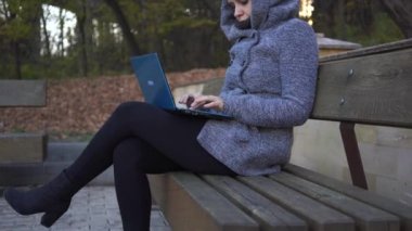 Uzakta bir dizüstü bilgisayarda sonbaharda bir bankta otururken yazarak sıcak bir kapüşonlu bir kadın. Kız parkta bankta laptop yazma