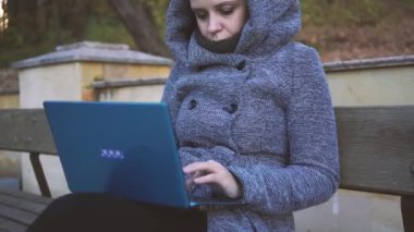 Uzakta bir dizüstü bilgisayarda sonbaharda bir bankta otururken yazarak sıcak bir kapüşonlu bir kadın. Kız parkta bankta laptop yazma