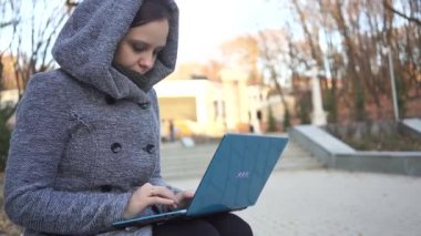 Uzakta bir dizüstü bilgisayarda sonbaharda bir bankta otururken yazarak sıcak bir kapüşonlu bir kadın. Kız parkta bankta laptop yazma