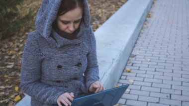 Uzakta bir dizüstü bilgisayarda sonbaharda bir bankta otururken yazarak sıcak bir kapüşonlu bir kadın. Kız parkta bankta laptop yazma