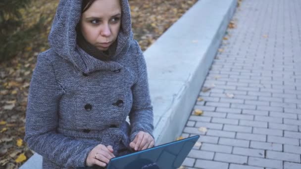 一名身穿温暖头套的女子坐在公园长椅上 在笔记本电脑上打字 女孩在笔记本电脑上写作在公园长凳上 — 图库视频影像