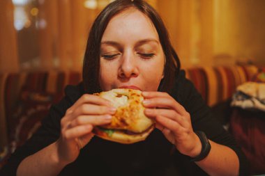 Kadın yemek hamburger Fransız. Öğrenci portresi lokanta masada tüketmek. Kız önemsiz yemek yiyor. Lokanta daek arka plan üzerinde reklam. Öğrenci yarı bitmiş ürünler yiyor.