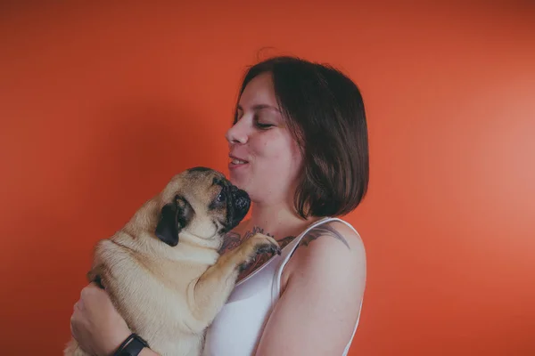 Beautiful girl with tattoo holding cute pug puppy on her hands, on orange background. The concept of friendship between man and dog.