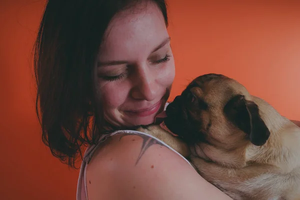 Beautiful girl with tattoo holding cute pug puppy on her hands, on orange background. The concept of friendship between man and dog.