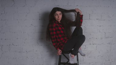 Kız kırmızı siyah ekose gömlekli. Esmer bir tuğla duvara bir sandalye ile poz. Gri arka plan üzerinde güzel kadın. 