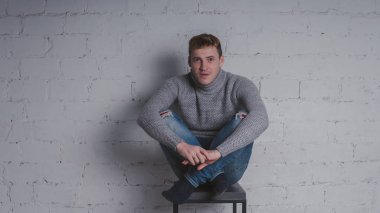 Sandalyeye gri bir arka plan üzerinde sıcak kazaklı adam. Bir adam bir tuğla duvara poz.