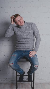 Sandalyeye gri bir arka plan üzerinde sıcak kazaklı adam. Bir adam bir tuğla duvara poz.