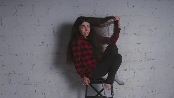 The girl in the red black plaid shirt. Brunette posing with a chair, against a brick wall. Beautiful woman on grey background.