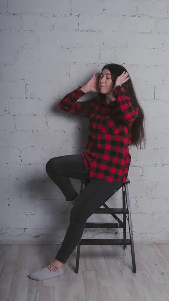 The girl in the red black plaid shirt. Brunette posing with a chair, against a brick wall. Beautiful woman on grey background.