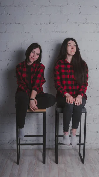 两个穿着红色和黑色格子衬衫的年轻女孩坐在砖墙背景的椅子上 女性友谊概念 — 图库照片