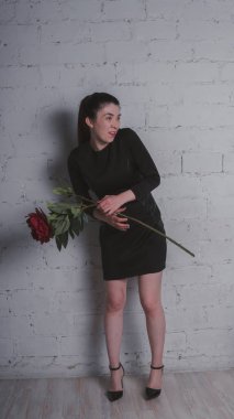 Bir ince kız siyah bir elbise içinde duran, büyük bir yapay çiçek tutan ve gri bir duvara poz