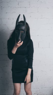 Siyah elbiseli, antik maskesi, stüdyoda bir beyaz tuğla duvara olan genç bir esmer fotoğraf