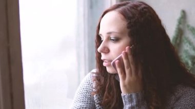 Güzel kız elinde smartphone ile pencere eşiğinde oturuyor. O uzun saçlı, gülümseme ve seyir onun telefon vardır.